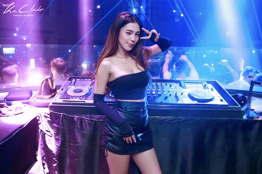 a Thai female dj posing in front of her booth while people are dancing on the background in the club khaosan