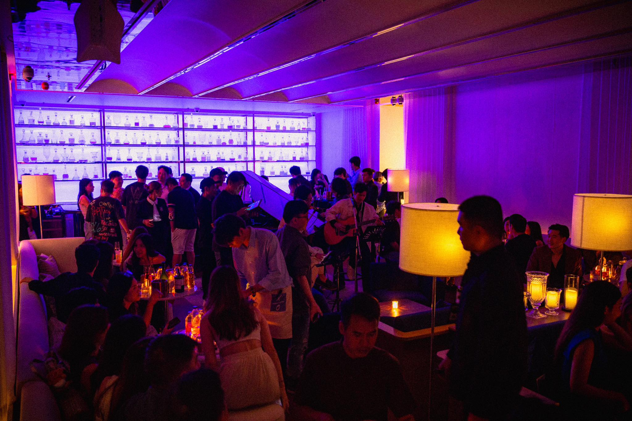 TIC TAC TOE is dimly lit with purple and orange lighting, creating a vibrant and lively atmosphere. There are numerous people gathered in the space, some standing and others seated. The bar area is visible, with shelves of liquor bottles and glasses behind it. The overall scene depicts a busy, social gathering in an upscale, nighttime venue.